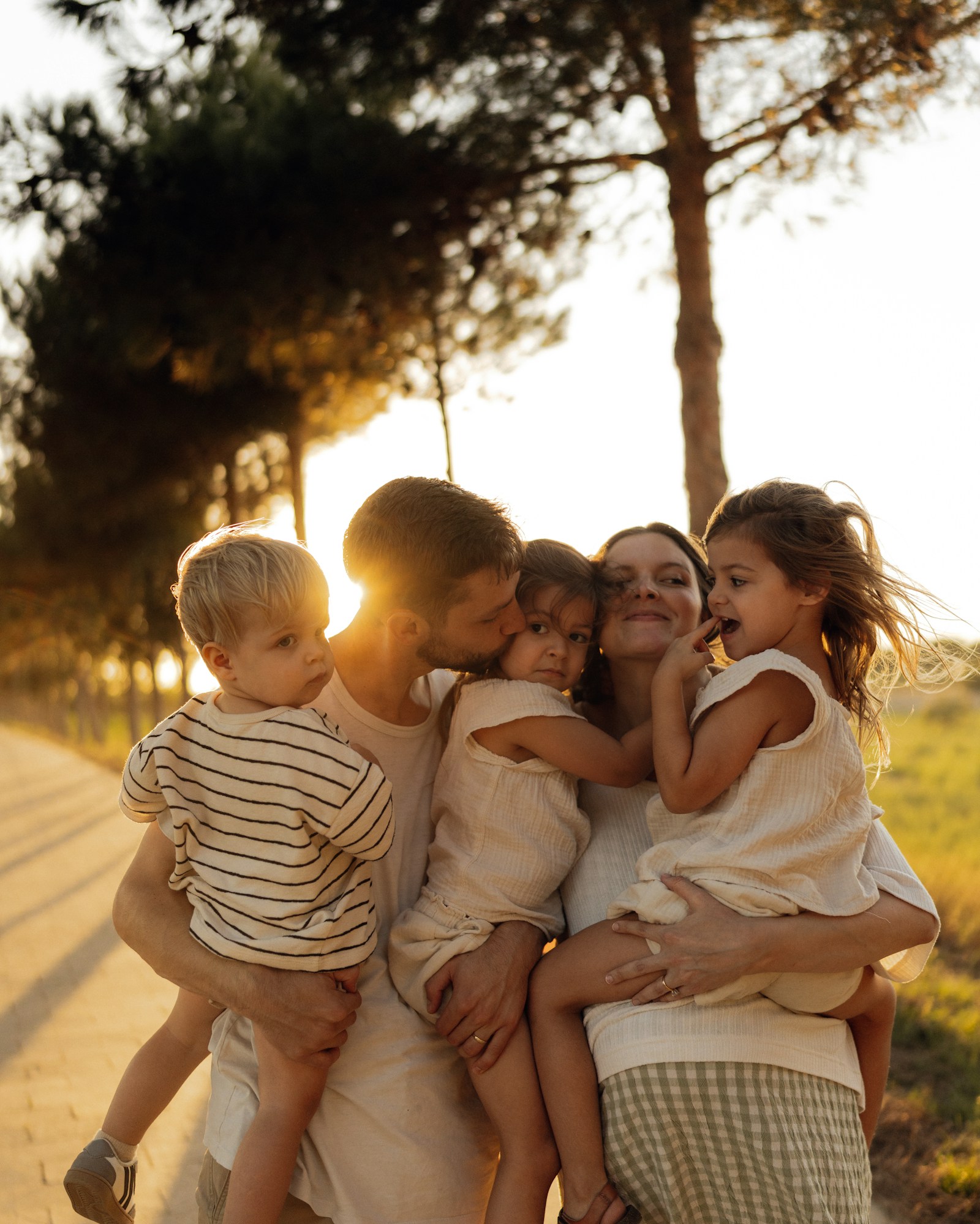A father and mother holding their three young children in their arms along a tree-lined path at golden hour